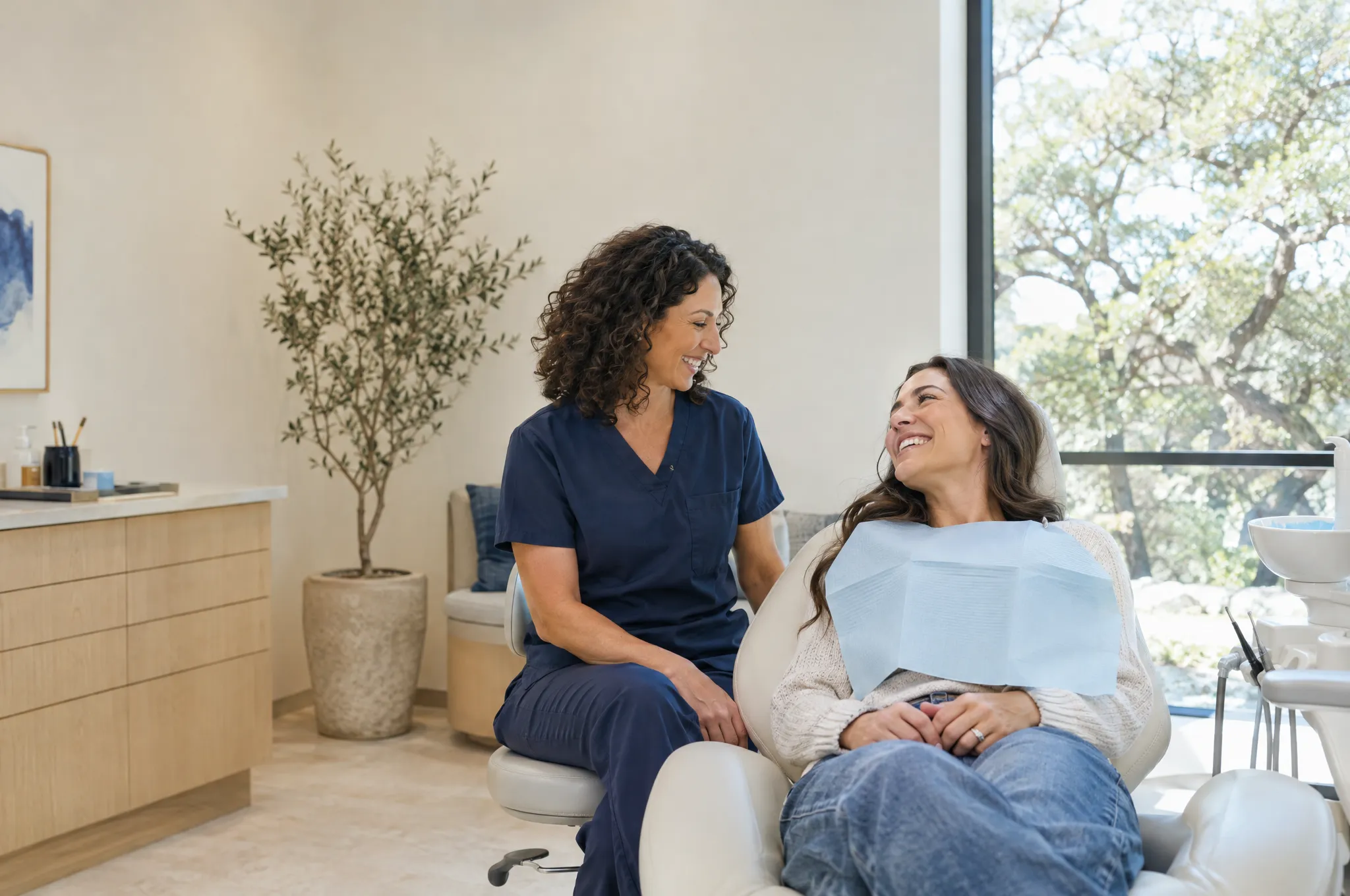 A dentist in navy scrubs laughing with a relaxed patient in a bright treatment room